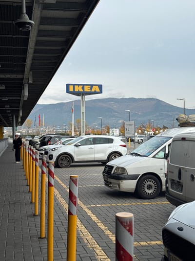 The scene depicts an outdoor parking lot and the exterior of an IKEA store in Sesto Fiorentino, Italy, under an overcast sky.
In the foreground, a dark grey paved walkway runs along the base of a substantial dark-colored roof structure extending from the upper left. A black dome-shaped surveillance camera is mounted beneath this roof. A row of yellow and red/white striped bollards separates the walkway from the parking area. One individual, dressed in dark clothing with white shoes and a white hat, stands on the walkway near the left edge, holding a white object to their head, consistent with using a mobile phone.
The midground features a paved parking lot with yellow demarcation lines. Several vehicles are parked: a white Kia Sportage SUV, an older white multi-purpose vehicle (possibly Fiat Scudo/Ulysse) with visible items inside, and the rear of a white van (Italian license plate "EC 103FE") with bundled items secured on its roof rack. In the center, a tall pole supports a large, triangular blue sign with yellow "IKEA" lettering. Below the sign, multiple flagpoles display flags, including one identifiable as the Romanian flag. Streetlights are visible across the parking lot.
The background reveals a range of mountains covered with sparse vegetation, positioned beneath a uniformly grey, cloudy sky. Buildings and structures are visible at the base of the mountains.