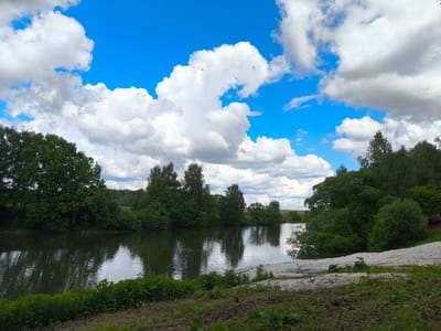 The image depicts a landscape scene in Mar'inskoe, Russia, featuring a body of water, possibly a pond or lake, surrounded by trees and vegetation. The sky is blue with numerous white, puffy clouds. The water reflects the sky and trees. The foreground is mostly green with grass and some bare earth. There are no visible people or animals in the image.