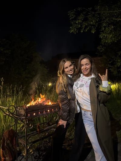Two young women are posing for a photo in the evening in a backyard setting. A bonfire burns in a decorative metal grill to their left, with smoke rising. Tall grass and trees form the background, with some outdoor lighting visible. The woman on the left is wearing a dark puffy jacket and has her arm around the woman on the right. The woman on the right is wearing a white hoodie with graphics, a dark olive coat, and light blue jeans. She is sticking her tongue out and making a rock-on hand gesture. The scene appears to be in the city of Савельево, Russia, based on contextual information.