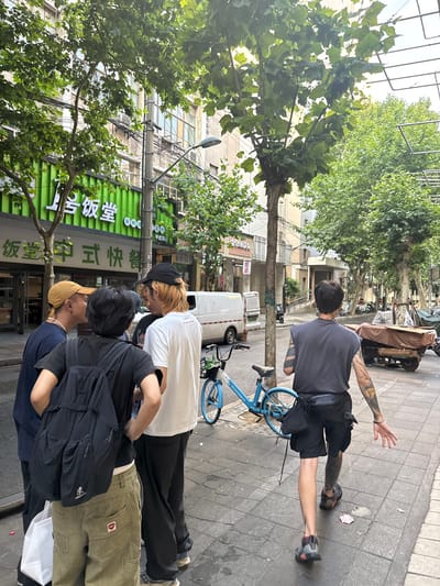 This daytime urban scene is situated on a paved sidewalk in Changning District, China, alongside a multi-story building featuring a prominent green facade sign displaying Chinese characters "上号饭堂" and "式快餐" (suggesting a canteen or fast food establishment). Five individuals are present. Four are grouped on the left-center of the frame: one in a yellow baseball cap and dark blue shirt with a white beaded necklace, another with a black backpack over a black t-shirt and olive green pants featuring a red lip-shaped patch on the rear pocket, and a third in a white t-shirt and black cap, exhibiting light-colored hair. A fourth individual is partially obscured within this group. They appear to be engaged in conversation. To the right, a fifth individual walks away from the group, viewed from behind. This person wears a dark grey sleeveless top, black shorts, a black waist bag, and dark athletic shoes, displaying extensive tattoos on both arms and the right calf. A blue shared bicycle is parked on the sidewalk near the group. The street is lined with mature trees, and a utility pole with a streetlight is visible. Parked vehicles include two white vans, multiple motorcycles, and a tarp-covered three-wheeled cargo vehicle further down the street. The sidewalk is tiled, and minor litter is visible.