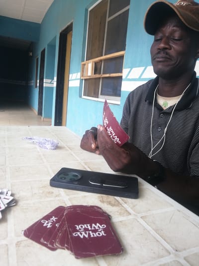 A middle-aged Black man wearing a brown baseball cap and a striped polo shirt sits at a white, plastic-covered table outside a building with light blue walls. The man has his eyes closed and is holding a red card with white text that appears to say "Whot" and "Who". His left hand is visible, resting on the table, with a ring on his finger. To his right, on the table, is a black smartphone with a camera array. In front of the man, a stack of red cards with white text, similar to the one he holds, is fanned out. To the left of the cards, there is a crumpled piece of translucent purple plastic. The background shows a hallway with doorways and windows. The scene is likely in the city of Ajasse Ipo, Nigeria, given the context provided.