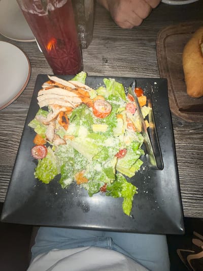 A top-down shot shows a dark-colored rectangular plate resting on a wooden-textured surface. The plate holds a salad featuring romaine lettuce, cherry tomatoes, croutons, sliced grilled chicken, and a sprinkle of shredded cheese. A pair of salad tongs rests on top of the salad.
To the left of the plate, there is a glass filled with a dark red liquid, along with a white plate. Above the plate, a wooden board with a bread item rests. To the right, a person wearing light blue jeans is partially visible.
The image was likely taken indoors at a restaurant. Further information is needed to confirm if it is located in Glenview, United States of America.