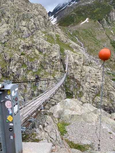 This image depicts the Trift Bridge, a pedestrian suspension bridge, located in a mountainous region near Innertkirchen, Switzerland. The scene is dominated by steep, rugged rock faces with sparse green vegetation and patches of snow on higher peaks in the background under an overcast sky. A strong stream or small waterfall flows down the right mountainside. The suspension bridge, constructed with a wooden deck and steel cables, stretches across a deep gorge, with one individual wearing a yellow backpack visible walking on it, approximately one-third of the way from the foreground. In the immediate foreground on the left, a metal support pillar for the bridge is heavily adorned with various stickers, including dates and logos. On the right side of the foreground, a large, bright orange spherical object is mounted on top of a tall metal pole, which is secured by cables anchored into the rock.