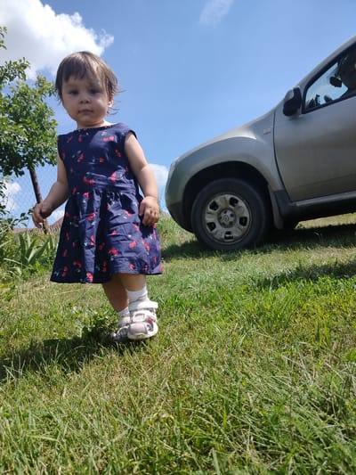 A young child stands on a grassy lawn in front of a silver car, under a blue sky with scattered clouds. The child, facing forward, is wearing a dark blue dress with a cherry pattern and white sandals. The car appears to be parked on an incline, with its front wheel visible. The surrounding area shows greenery, including grass and some foliage. The scene suggests an outdoor, daytime setting, possibly a residential area or a park. The specific location is Блохино, Russia.