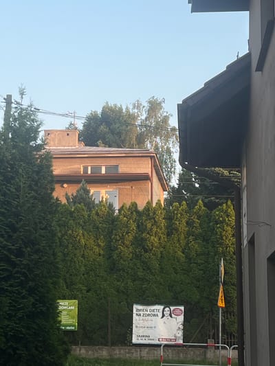 The image shows an outdoor scene on a clear day with a bright blue sky. In the foreground, a dense row of green evergreen trees lines a fence. To the left, a green billboard is partially visible, displaying text in white. To the right of the green billboard, a larger white billboard features an image of a woman and text in Polish. A street sign with a bicycle symbol is mounted on a pole behind the white billboard. In the background, a two-story building with a light-brown facade and a dark roof is visible, partially obscured by trees. The building has windows and a chimney. Another building, a darker structure, is on the far right of the image, extending from the bottom to the top. Electrical wires are strung across the sky, supported by a utility pole on the left. The location is identified as Skawina, Poland, as indicated by text on the white billboard.