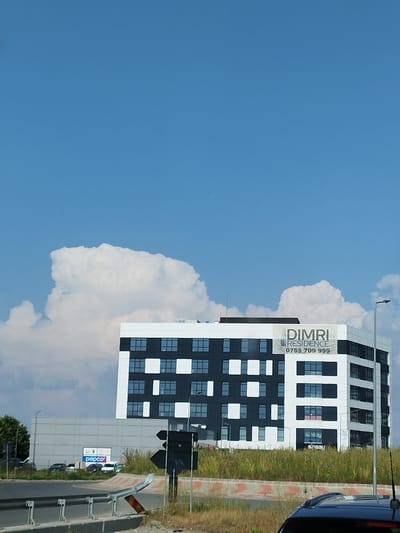 The image depicts the DIMRI RESIDENCE building, a modern, multi-story structure with a facade featuring alternating black and white panels and numerous windows. A banner on the building displays "DIMRI RESIDENCE" and a phone number, "0755 709 999." Adjacent to DIMRI RESIDENCE is a lower, grey building with a "pepco" sign. The scene is set against a bright blue sky with scattered white clouds. In the foreground, a road curves to the left, bordered by a metal guardrail. The general environment appears to be an urban or semi-urban area, likely on the outskirts of a city, with vegetation growing along the roadside. The image captures a daytime scene under clear weather conditions. The location is identified as Bragadiru, Romania.