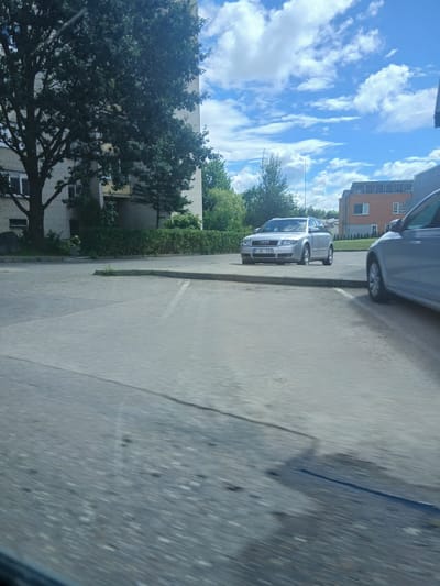 This is a slightly blurry, eye-level, outdoor photograph taken on a sunny day in Garliava, Lithuania. The scene depicts a parking lot with two cars. On the left, a silver Audi A4 Avant is parked. Its license plate reads "FJK 159". To its right, partially visible and out of focus, is the side of another silver car. The background features a light-colored brick building with trees and hedges in front of it, and a modern orange brick building with solar panels on its roof further in the distance. The sky is blue with fluffy white clouds. The foreground is a paved asphalt surface, showing some cracks and a dark rectangular patch, possibly indicating a repair or stain.