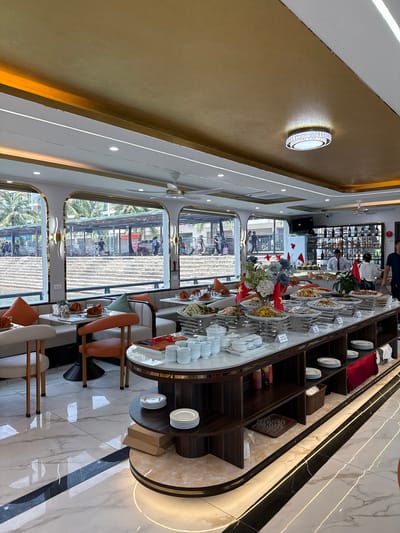 This image captures the interior of a luxurious dining area, likely a restaurant or a buffet hall, possibly on a cruise ship or overlooking a waterfront in Tuan Chau Ward, Vietnam. The setting is modern and elegant, featuring a golden-textured wall on one side, white walls with sleek gold trim, and a glossy marble-patterned floor accented with black and gold stripes.

A long, curved buffet counter dominates the central space, meticulously arranged with numerous dishes, stacked white plates, bowls, and serving utensils. Small Vietnamese flags and floral arrangements (featuring blue and white flowers) are interspersed among the food displays. Several people are visible: staff members in uniform attend to the buffet, and patrons are seated at dining tables with light and orange upholstered chairs, or moving about the area.

Large arched windows line the opposite side of the room, offering a bright view of the outdoors. Through these windows, a vibrant scene unfolds, showing many people walking along what appears to be a pier, boardwalk, or a stepped pathway. Palm trees and other outdoor structures are also visible, suggesting a sunny daytime setting. Decorative elements include a circular wall light with sparkling details, recessed ceiling lights, and ceiling fans. The overall ambiance is refined and organized, indicating an upscale dining experience. No legible text is clearly visible on any signs or objects within the image, beyond unreadable characters on small buffet labels.