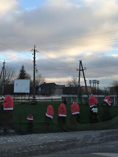 The image depicts a street scene in Illintsi, Ukraine, under a partially cloudy sky with hints of blue. In the foreground, a dark gray paved surface is visible, reflecting some light. Immediately beyond this, a green grassy area features a linear arrangement of six dark green bushes or small evergreen trees. Each of these six bushes is topped with a red and white Santa hat; the fifth bush from the left (second from the right) also incorporates a white, fluffy ornament and a small red object below its hat, while the sixth bush (far right) wears a red and white hat with a floral pattern. A smaller bush, located slightly forward and to the left of the main row, is topped with a smaller red and white Santa hat.
In the middle ground, from left to right, a utility pole with multiple power lines is present. To its right, a prominent, tall evergreen tree stands. Further right, another dark utility pole supports numerous power lines crisscrossing the upper frame. Adjacent to this pole, a large, blank white rectangular sign is mounted on a separate pole. To the right of the blank sign, a blue square road sign with a white pedestrian crossing symbol is visible. Behind these elements, various bare deciduous trees and several residential buildings with dark, sloped roofs are discernible, partially obscured by a light-colored fence line. In the far background, on the right side of the frame, four light-colored, cylindrical water towers stand against the horizon. Multiple black power lines traverse the upper portion of the scene against the sky. No individuals are present in the image.