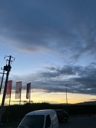 The image captures a dramatic outdoor scene at either sunrise or sunset, characterized by a dynamic sky and strong contrasts of light and shadow. The upper and left portions of the sky are filled with heavy, dark blue-grey clouds, which gradually lighten and become more textured towards the lower and right side of the frame, where a brilliant golden-orange glow marks the horizon.
In the foreground on the left, a utility pole with multiple insulators and wires is silhouetted against the sky. To its right, three red banner flags are visible on poles, displaying inverted Arabic text that is not clearly legible. Further right, a slender street light with a curved arm extends upwards.
The horizon is defined by a vivid band of yellow and orange light, suggesting a vast, open landscape or distant body of water. Below this luminous horizon, dark silhouettes of vegetation, possibly bushes or trees, are present. On the far right, parts of parked vehicles are visible, including the front section of a white van and a darker car positioned behind it.
The scene is static, with no discernible human activity, and the overall impression is one of serene, atmospheric beauty. The visible elements, including the Arabic script on the flags, align with the location context of Ain Kansra, Morocco.