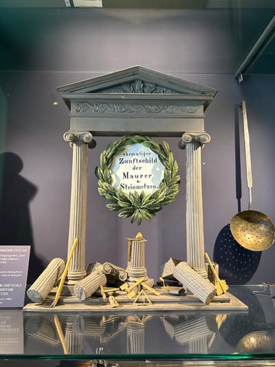 The image shows a Zunftschild (guild sign) for stonemasons and bricklayers. It is an artifact displayed in a glass cabinet. The sign is composed of classical architectural elements, including columns, a pediment, and a laurel wreath surrounding a central inscription in German: "ehemaliger Zunftschild der Maurer & Steinmetzen" (former guild sign of bricklayers & stonemasons). The sign also includes broken columns and tools, suggesting labor and potentially construction or destruction. Above the sign, a metal skimmer with holes is visible. In the bottom of the frame there is a table with the title of what is inside the cabinet: STUTZUHR.

The display includes printed labels with additional information. A partial label is visible with the text "RMACHER: STUTZUHR" and some text in the German language relating it to the city of Biberach an der Riß.