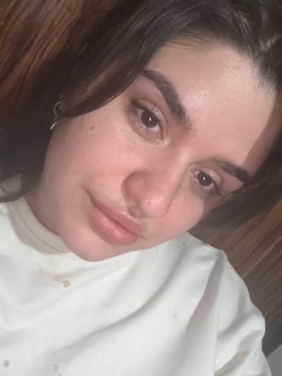A close-up, high-angle shot shows a young person's face, cropped from the chest up, against a dark wooden background. The individual has dark hair, thick eyebrows, and brown eyes. Their lips are slightly parted, revealing a subtle smile. They are wearing a white t-shirt with what appear to be light brown stains on the fabric. The lighting is warm and diffused, casting soft shadows. The scene does not provide any specific information about its location in Kadiivka, Ukraine.
