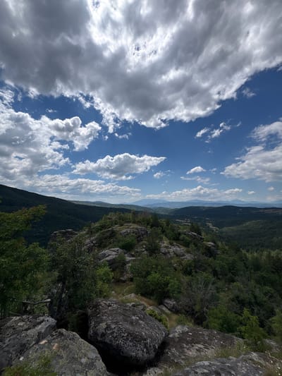 This is a landscape photograph taken from a rocky, elevated viewpoint overlooking a valley and distant mountains. The foreground is dominated by large, grey, moss-covered rocks and sparse green vegetation. The middle ground features a densely forested hillside on the left, with a less dense, more varied green landscape stretching into the valley. In the distance, layers of rolling hills and mountains fade into a hazy blue horizon. The sky is a vibrant blue, filled with a dramatic display of white and grey cumulus clouds. The scene appears to be in Dolno Drianovo, Bulgaria, capturing a natural, mountainous environment under a partly cloudy sky. There are no people or identifiable structures in the frame.