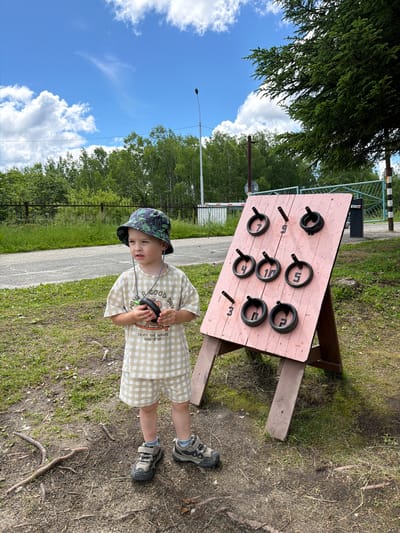A young boy stands in a grassy area next to a ring toss game in Kul'dur, Russia. The boy, wearing a bucket hat and a checkered t-shirt and shorts, holds a black ring in his hands. He is looking to his right. The ring toss game is set up on a wooden easel and has a pink board with pegs and rings attached. The background features trees, a road, and a blue sky with white clouds. A metal fence and a gate are visible in the distance.