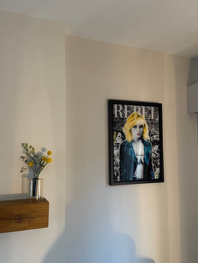 The image captures an indoor scene dominated by light-colored walls with minimalist decor. A rectangular framed artwork is mounted on the upper portion of the wall. This artwork features a pop-art style portrait of a blonde woman with red lipstick, wearing a blue leather-like jacket over a light-colored shirt. The background of the portrait is a collage of black and white patterns and smaller images. Visible text within the artwork includes "BLONDIE" printed vertically along the left edge, and "UNDER" partially visible at the bottom left. Another phrase, possibly "my word," can be faintly seen on the woman's shirt.

Below the artwork, on a lower wall section, a small arrangement of artificial yellow flowers with green leaves is held in a clear, cylindrical glass vase. This vase is inserted into a sturdy, light-brown wooden block or beam, which is affixed to the wall. Shadows from the flowers and vase are cast onto the wall to the right. The walls themselves are a uniform, light off-white or cream color, with subtle tonal variations suggesting ambient lighting. The scene appears calm and well-lit, typical of a modern interior. The setting for this media file is Harrogate, United Kingdom.