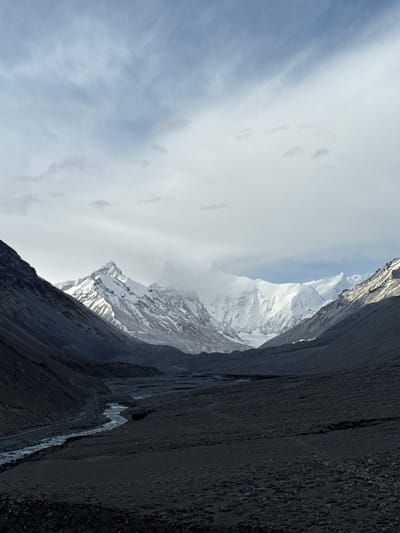 The image depicts a stark mountain landscape in Tashi Dzom town, China. A snow-capped mountain pierces through a cloudy sky, partially obscured by mist. To the right, a steep, dark cliff face dominates the foreground, exhibiting a coarse, rocky texture. A narrow stream or drainage channel runs vertically along the cliff. The sky is pale blue with wisps of white clouds to the left of the mountain.