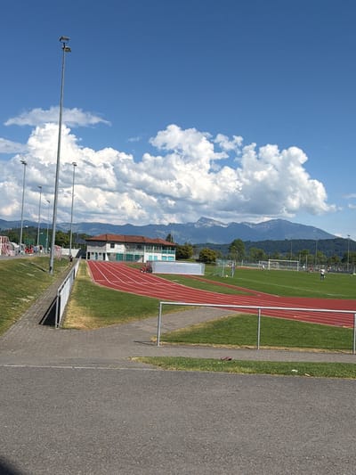 The image depicts a track and field stadium in Rapperswil-Jona, Switzerland. A red running track curves around a green field, with athletic equipment visible in the background. A building with multiple windows and a red roof is adjacent to the track. Beyond the stadium, mountains are visible against a blue sky with fluffy white clouds. Street lights line the foreground, and a paved pathway runs along the edge of the stadium. People are visible near the soccer field.