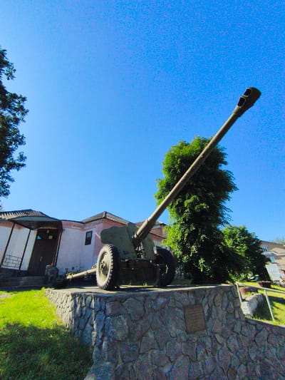The image displays an outdoor scene in Velyka Bahachka, Ukraine, featuring a large, olive-green artillery piece, likely a field gun or howitzer, mounted on a stone pedestal. The cannon's long barrel extends diagonally upwards and to the right, pointing towards the clear, bright blue sky. Its muzzle is distinctive with two large, dark openings. The artillery piece has large, spoked wheels with rubber tires.

The pedestal supporting the cannon is constructed from irregularly shaped, rough-hewn grey-brown stones. A rectangular plaque is visible on the front face of the pedestal, but the text engraved on it is illegible.

In the background to the left, a building with light-colored, possibly pinkish-beige, walls and a dark, ridged roof is visible. It features an entrance with a dark door and an overhanging canopy. On the far left, the foliage of a large tree borders the sky. To the right of the cannon, a tall, dense green tree stands, its vibrant foliage partially framed by the cannon's barrel. Further in the background on the right, another building and a sign with unreadable text are faintly visible.

The ground around the monument is covered in lush green grass, with bright sunshine casting clear light and shadows, indicating a daytime setting under fair weather conditions. No people are visible in the scene. The overall impression is of a public memorial or outdoor exhibit.