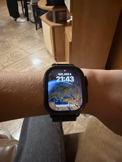 A close-up image shows a person's left wrist wearing a black Apple Watch Ultra. The watch face displays a world map with the time "21:43" and the date "SAT, APR 4." The humidity is listed as "86%." The wrist is fair-skinned with visible hairs. The background includes portions of a wooden table, floor tiles, and a person's gray pants and white shoes. No specific location is identifiable from this image.