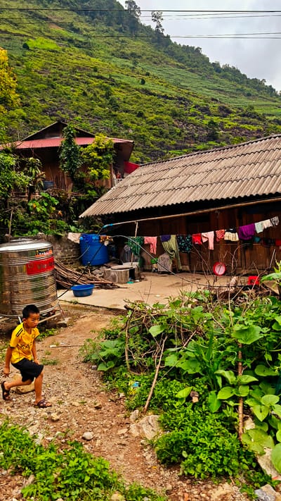 A young boy in a yellow shirt and black shorts walks on a rocky, dirt path in the Cán Tỷ Commune, Vietnam. He is wearing brown sandals. To the right of the path, lush green vegetation grows. In the background, a house with a corrugated tile roof and clothes hanging on a line in front is visible. Next to the house is a stainless steel water tank, a blue plastic water tank, and a stack of firewood. Another house is visible further up the hillside. The background is a steep, green, tree-covered mountain. The sky is overcast.