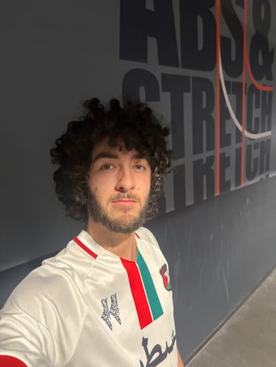 A person with curly hair and a beard lies on a gray mat with large black text "STRETCH" repeated across it. The person wears a white shirt with a red and green stripe on the shoulder and a stylized symbol on the chest. The shirt is long-sleeved and appears to be a sportswear or team jersey style. The image is taken at an angle, possibly a selfie. The lighting is soft and even.