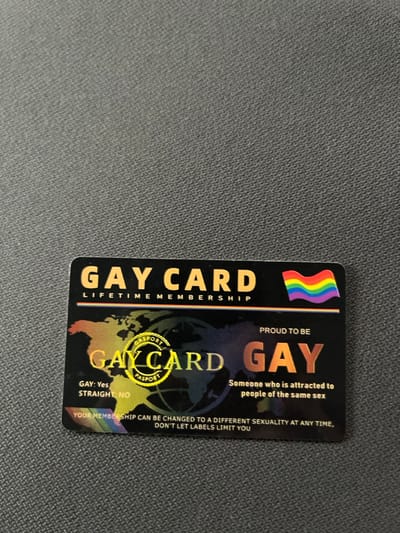 A black, rectangular novelty card, labeled "GAY CARD LIFETIME MEMBERSHIP," is resting on a dark grey, textured fabric surface in Maslarevo, Bulgaria. The card features a rainbow flag emblem in the upper right corner. Below the main title, a stylized holographic world map is present, overlaid by a circular yellow seal marked "GASPORT PASPORT." Textual elements on the card include "GAY: Yes" and "STRAIGHT: NO" on the left side. On the right, "PROUD TO BE GAY" is displayed, followed by the definition "Someone who is attracted to people of the same sex." The bottom of the card contains a statement: "YOUR MEMBERSHIP CAN BE CHANGED TO A DIFFERENT SEXUALITY AT ANY TIME, DON'T LET LABELS LIMIT YOU," presented over a subtle rainbow gradient. No individuals or actions are depicted in the scene.