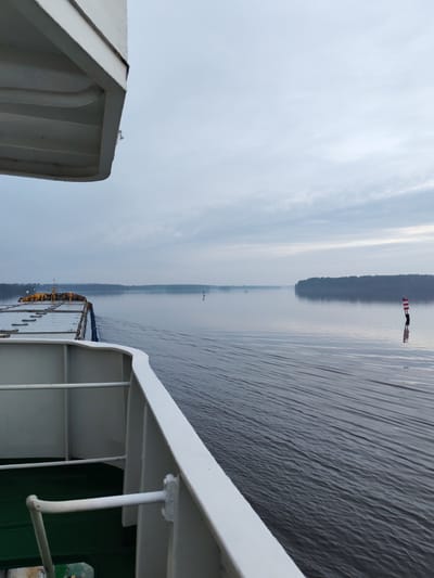 The image captures a view from the deck of a cargo ship navigating a wide, calm body of water, likely a river or lake, under an overcast sky. The ship's white railings and superstructure are prominent in the foreground, partially obscuring the view of the cargo area which is covered with tarpaulins. The water shows a gentle wake trailing behind the ship. In the distance, a treeline borders the opposite shore, suggesting a natural, possibly rural, environment. A red and white buoy marks a channel in the water, and a few other smaller buoys are visible further out. The overall atmosphere is subdued, hinting at either early morning or late evening light. There are no people visible in the frame. Based on the visual cues, the location is consistent with the waterways near the settlement of Shindalovo, Arkhangelsk Oblast, Russia.