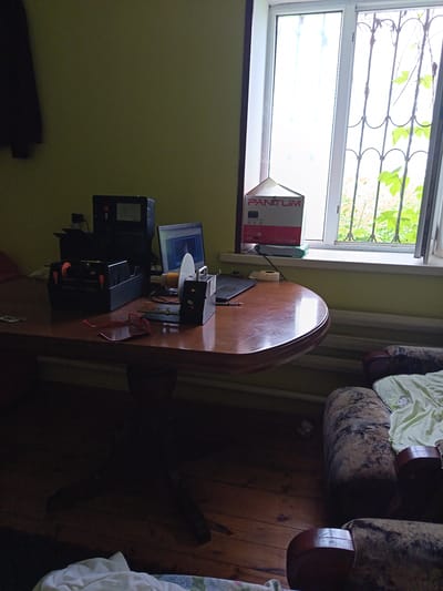 A dimly lit room features a wooden table laden with office equipment, including a laptop, a printer, and a label maker. A pair of red-rimmed glasses rests on the table. To the right, a window with bars overlooks a verdant, overgrown outdoor space, suggesting a possibly warm day. A Pantum printer box sits on the windowsill. The room appears to be a workspace, possibly in a home, with a couch visible in the foreground. The overall mood is somewhat cluttered and domestic. The time of day is unclear due to the interior lighting, but the brightness from the window suggests daylight.