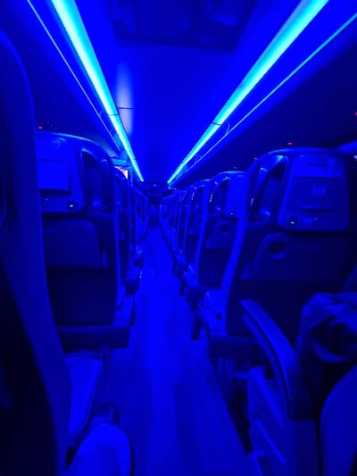The image displays the interior of a passenger transport vehicle, likely a coach or bus, characterized by intense blue ambient lighting. Two parallel strips of bright blue LED lights illuminate the ceiling and cast a strong blue hue across the entire cabin. Rows of unoccupied, dark-colored passenger seats with integrated headrests and visible seat-back components, possibly infotainment screens or charging ports, line both sides of a central aisle. The aisle floor is also bathed in blue light and extends into the background, showing additional rows of seating in diminishing perspective. No individuals are visible within the frame, and no specific actions or interactions are depicted. The environment is orderly and appears to be in operational condition, possibly awaiting passengers or traveling at night. The specific location within any city in South Oxfordshire, United Kingdom, cannot be determined from the visual information provided.