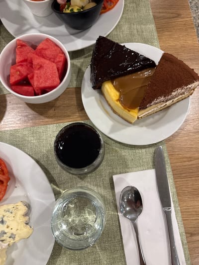 The image shows a table setting at a restaurant in Es Camp De Mar, Spain. On the table are several plates of food and drinks. One plate contains three slices of cake: a chocolate cake, a caramel cheesecake, and a tiramisu. Another plate has watermelon slices. A third plate holds a slice of cheese and a slice of tomato. There are also glasses of water and dark liquid, a spoon, and a knife. The table is covered with a green placemat, and the background is a wooden surface. The overall scene suggests a meal or dessert being served at a restaurant.