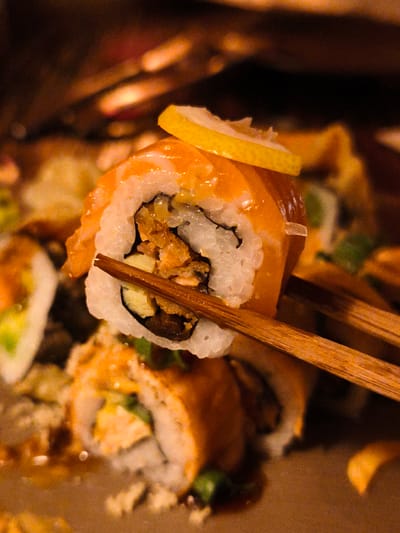 A close-up, slightly blurred, vertical shot captures a piece of sushi being held by wooden chopsticks. The sushi roll features a bright orange slice of salmon on the outside, encasing rice, and various fillings visible within. A thin slice of lemon rests on top of the salmon. In the background, other sushi pieces and blurred elements of a dish are visible, with warm, ambient lighting creating a cozy, indoor setting, possibly a restaurant. The focus is sharp on the sushi piece and chopsticks, highlighting the textures and colors of the food. The overall mood is appetizing and inviting. No text is visible. The location is Be'er Sheva, Israel.