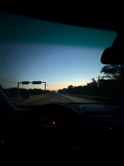 The image depicts a view from inside a car driving on a highway at dusk. The sky is a gradient of deep blue to orange as the sun sets. Trees line the sides of the highway, and a large overhead road sign with two rectangular panels is visible in the distance. The dashboard of the car is dark, with illuminated red gauges showing speed and other information. The overall atmosphere suggests a peaceful journey as daylight fades. The location is identified as Voyvodinovo, Bulgaria.
