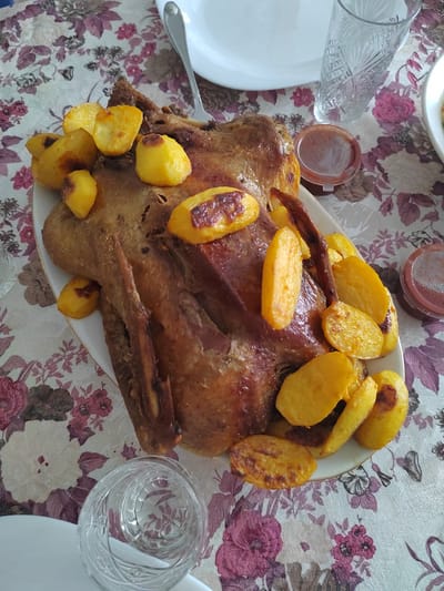 Here's a detailed description of the image:

**Scene:** An indoor setting, likely a dining area or kitchen, judging by the tablecloth and dishes.

**Subjects:** A roasted duck (or similar fowl) dominates the center of the frame. It is plated and surrounded by roasted potatoes. There are also small containers of sauce nearby.

**Objects:**
*   A floral patterned tablecloth is spread across the table.
*   A white plate holds the roasted duck and potatoes.
*   A glass and other plates are on the table.
*   Silverware (a spoon or fork) is visible.
*   Small containers with sauce.

**Actions/Interactions:** The duck has been cooked and plated for consumption. The potatoes are arranged around it.

**Environment:** The image is well-lit.

**Location (Италмас, Russia):** There is nothing in the image to confirm that this image was taken in Италмас, Russia.
