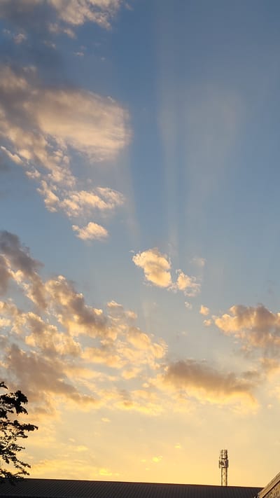 This media file depicts a dramatic sunset sky over Hengersberg, Germany. The scene is dominated by a vast expanse of sky, with fluffy, golden-tinged clouds scattered across a fading blue canvas. The clouds are illuminated by the setting sun, casting warm hues of orange and yellow, contrasting with the cooler blue of the upper atmosphere. Faint sunbeams are visible, adding depth and texture to the sky.
On the right side of the frame, a silhouette of a building with a textured roofline suggests an urban or industrial setting. Rising above this, a section of a construction crane is visible against the bright sky, hinting at ongoing development or infrastructure work in the area. At the very bottom right, the dark silhouette of tree branches adds a natural element to the scene, framing the lower edge of the composition.
The time of day is clearly late afternoon or early evening, given the intense golden light and the sunset colors. The weather appears clear, with the clouds indicating typical atmospheric conditions for a sunset. There are no people visible in the image, and no text is present. The overall mood conveyed is one of natural beauty and transition, as day gives way to night.