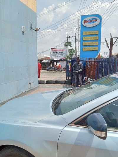 The image is taken from a street corner in Sapele, Nigeria, located at latitude 5.878 and longitude 5.699. A silver car is parked on the street corner. In the background, there are several people on foot, some are riding bicycles, and others are walking. A large sign in blue and white advertising Memories Superstore is in the upper right corner of the image. The sign lists various products sold at the store including foods, beverages, ice cream, household goods, and pharmaceuticals. A sign for Living Faith Church is visible on a building in the background. The image captures a typical street scene in Sapele, Nigeria, highlighting the presence of businesses and pedestrians.