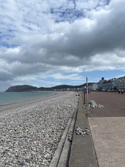 The photograph depicts a pebbled beach with a concrete promenade in Llandudno Junction, United Kingdom. The beach is predominantly covered with grey pebbles. The promenade is constructed with concrete. In the background, there are buildings and a hillside visible under a cloudy sky. The image is oriented vertically.