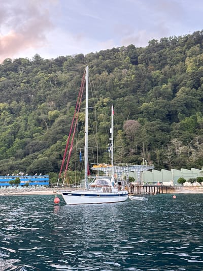 A sailboat is anchored in the water, its hull white with a blue stripe. The sailboat has a tall mast with rigging and flags. In the background, a lush green hillside covered in trees rises steeply. Along the shoreline, there is a blue building with multiple windows and satellite dishes, as well as a dock with industrial-looking structures. The water is dark blue with whitecaps and foam. The sky above is a soft pink and blue, suggesting either sunrise or sunset. The scene appears to be located on Christmas Island, Australia, a territory known for its tropical rainforests and coastal areas.