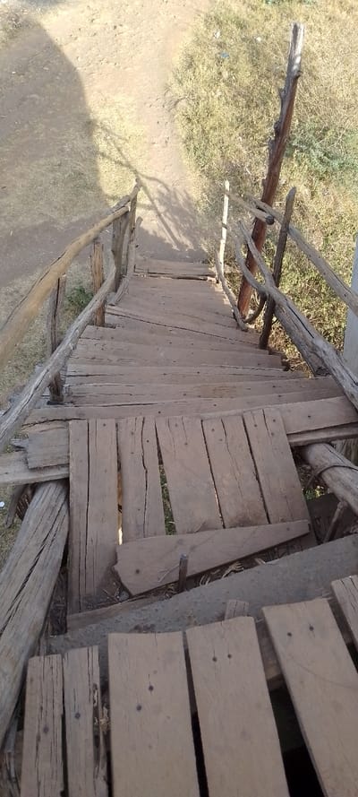The image presents a downward-looking view of a rustic, handmade wooden staircase or pathway situated outdoors. The steps are constructed from rough, uneven wooden planks, many of which show signs of age, wear, and weathering, including cracks and splinters. Visible nails secure the planks, some appearing to be partially raised.

The staircase features handrails on both sides, fashioned from natural, unrefined tree branches or small logs, which are held in place by vertical posts made from similar materials. The construction of both the steps and the handrails appears informal and possibly improvised, lacking standard finishes or precise alignment.

Below and to the left of the stairs, the ground is composed of light brown, dry earth, possibly a dirt path or cleared area. To the right, sparse, dry, yellowish-green grass and some low-lying shrubbery are visible. Shadows are cast across the ground and some parts of the steps, indicating bright daylight and a clear sky, likely during the morning or afternoon.

Notable details include significant gaps between several planks, particularly in the lower-middle section of the staircase, through which the ground and vegetation below can be seen. One large plank in the mid-ground appears slightly dislodged or tilted, further emphasizing the informal and possibly precarious nature of the structure. No specific activity is occurring, and there is no visible text in the image. The overall impression is that of a functional but roughly built outdoor passage in a natural or undeveloped setting.