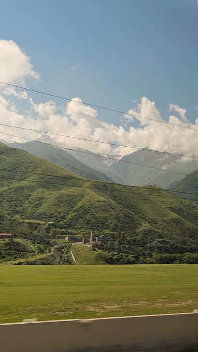 In Nizhnyaya Unal, Russia, a bright, overcast day provides a backdrop of a vast, blue sky dotted with fluffy white clouds. Dominating the scene are immense, lush green mountains, their slopes covered in dense vegetation. The mountains are layered, with the closest peaks a vibrant green, gradually fading into softer hues as they recede into the distance. Some of the higher mountain peaks are capped with snow, stark white against the green and blue.

Nestled on the slopes of the mountains is a small village. Several stone buildings, some appearing to be ancient towers, are visible, clustered together. Smaller residential buildings and scattered trees surround the village. A winding dirt path or road leads up the hillside towards the settlement.

In the foreground, a vast, flat expanse of perfectly manicured green grass stretches across the image. The grass appears to be a field, possibly for agriculture or recreation. Running across the bottom of the frame is a blurred, horizontal dark gray strip, suggesting the image was taken from a moving vehicle, likely a car, with a thick border or barrier in the immediate foreground. Several dark, thin lines, possibly power or telephone wires, stretch horizontally across the sky and over the landscape, indicating the presence of infrastructure. The overall impression is one of natural beauty, with signs of human habitation integrated into the mountainous terrain.