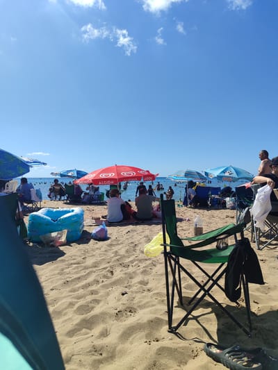 The image depicts a busy beach scene in Kandıra, Turkey, on a sunny day. The foreground is dominated by sand with several beach chairs, some occupied and some empty. A green folding chair with a black garment draped over it is prominently featured in the lower right. An inflatable blue kiddie pool is also present in the sand, along with various bags and items.
In the mid-ground, several people are seated under umbrellas, enjoying the beach. The umbrellas, some red with "ALGIDA" branding and others blue with patterns, provide shade. Further in the background, the sea is filled with swimmers, enjoying the water under a clear blue sky with scattered white clouds. The sun is bright, casting shadows on the sand. The overall atmosphere suggests a popular tourist destination during peak season.