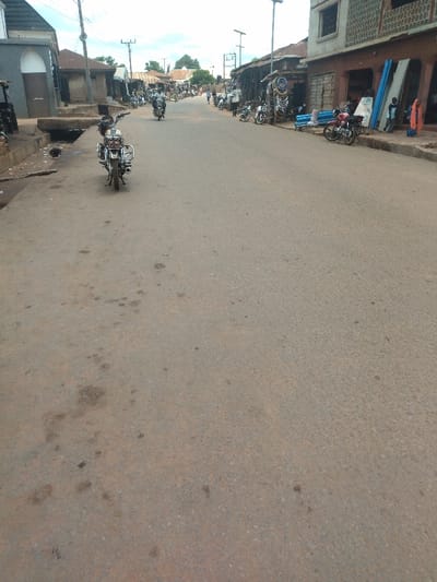 The image shows a street in Lokoja, Nigeria, taken from a point on the street looking down the length of the road. The coordinates indicate the location is in the city of Lokoja. The street is paved with asphalt and is relatively empty of traffic. There are two motorcycles parked on the right side of the street, and several more motorcycles are parked further down the road. There are also a few buildings along the street, all of which are single-story and appear to be commercial in nature. There are no people in the image. The sky is cloudy, and there are trees on either side of the street.  The asphalt road shows some wear and tear, with several noticeable dirt patches, and the road is in generally rough condition.