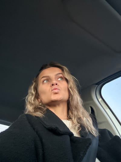 A fair-skinned woman with curly blonde hair and green eyes poses with pursed lips. She is wearing a white sweater and a black coat. The picture is taken from a low angle, looking up at her. The background shows the interior of a car, with the roof visible in the upper left and the top of the windshield in the upper right. The lighting is soft and diffused.
