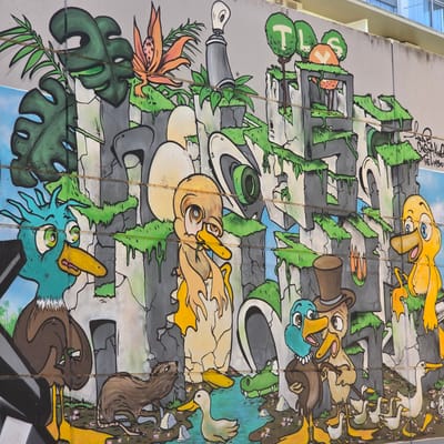 The image shows a section of a large, colorful mural on a building in Guardamar del Segura, Spain. The mural features cartoon-like depictions of ducks in various settings. Some ducks are wearing hats or engaging in human-like activities. The background includes representations of plants, trees, rocks, and water. The artwork is spread across multiple sections of the building's exterior, with lines dividing the panels. The style is vibrant and whimsical, suggesting a playful or educational theme.