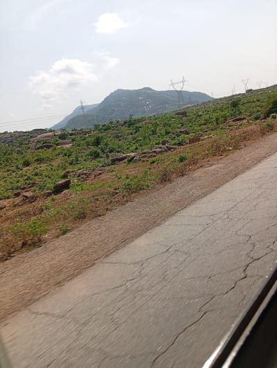 The image captures a scenic view of a hilly, rocky landscape dotted with green vegetation. In the background, large mountains rise under a bright, hazy sky with scattered clouds. Several tall, metallic electricity pylons are visible across the hillside, connected by thick power lines, indicating a developed area within the natural setting. A paved road, showing signs of wear and cracks, cuts diagonally across the foreground and mid-ground, suggesting it is a route through this terrain. The perspective seems to be from a moving vehicle, as indicated by the motion blur on the road and the slight tilt of the image. The terrain is rugged, with numerous large rocks interspersed with shrubs and trees. The overall impression is one of a rural or semi-rural area with infrastructure present. The time of day appears to be daytime, possibly late morning or early afternoon, given the brightness of the sky. There is no visible text in the image.
