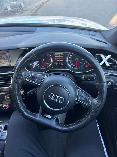 A car's interior is visible, including the steering wheel, dashboard, and center console. The steering wheel has an Audi logo in the center and the S Line badge on one spoke. The instrument cluster shows gauges and a digital display with some information, including "SD GALENA & MEDI" and a speedometer reading. Parts of the center console and dashboard show buttons, vents, and a display screen. A person wearing black pants is seated in the driver's seat, partially visible. The image does not provide enough information to identify the location as Dolni Tsibar, Bulgaria.