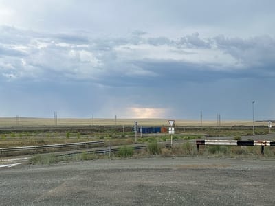An overcast sky with scattered clouds looms over a vast, flat landscape in Bozshakol, Kazakhstan. In the middle ground, a road appears to be under construction or undergoing maintenance, with a metal barrier painted in black and white stripes on the right. Further into the scene, a series of shipping containers, some blue and orange, are visible. A yield sign and other road signs are present. Power lines stretch across the horizon, punctuated by a few sparse trees and vegetation. The foreground consists of a gravelly surface. The lighting suggests either dawn or dusk, or a heavily overcast day with a break in the clouds in the distance.