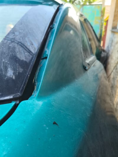 The image shows a close-up, angled view of the exterior of a teal-colored car, focusing on the area where the rear windshield meets the body. The car appears to be parked outdoors, possibly in a residential area or near a building with a concrete wall visible to the right. The car's surface is dusty and shows signs of wear, including a small dark object, possibly a dead insect, on the hood. The background is blurred, but suggests a fence or gate and greenery, consistent with an outdoor setting in Barysaw, Belarus. There are no people or ongoing activities depicted in the image.