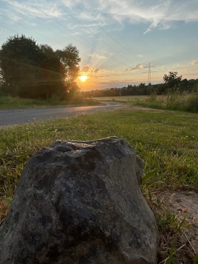 An outdoor, low-angle shot captures a large, dark grey boulder in the foreground, occupying the lower left portion of the frame. The boulder has a rough, textured surface and is partially covered by blades of green grass and dry straw. Behind the boulder, a paved path curves from the left and disappears into the middle ground. The path is flanked by green grass dotted with small white wildflowers. In the mid-ground and background, trees and fields stretch towards a horizon under a sky filled with dramatic clouds. The sun is setting, casting warm, golden light through the trees and creating sunbeams that fan across the sky. Power lines are visible strung between tall pylons in the distance. The environment suggests a rural or semi-rural setting, possibly on the outskirts of Neukirchen, Germany, given the presence of agricultural fields, trees, and infrastructure like power lines, typical of the German countryside. No people are visible in the image, and no actions or interactions are depicted.