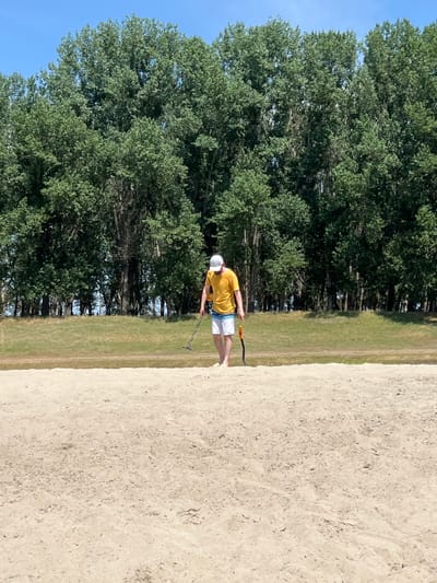 A man wearing a yellow t-shirt, white cap, and patterned shorts is metal detecting on a sandy expanse. He is walking from left to right across the frame, holding a metal detector and a shovel. Behind him, a line of tall green trees forms a dense backdrop under a clear blue sky. The ground in the foreground is a light-colored sand, and a grassy embankment is visible between the sand and the trees. The scene appears to be outdoors, possibly in a park or a natural area.