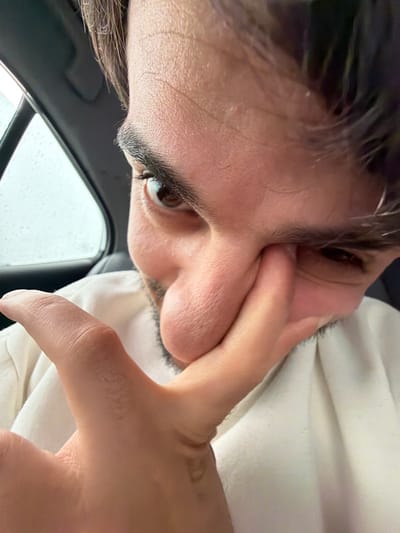 A close-up, low-angle perspective image focuses on a person's face. The person's hand is positioned against their face. A partially visible car window suggests an interior setting, and rain is visible on the window. The location is impossible to determine from the image alone.